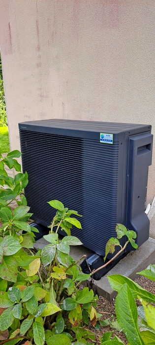 Unité extérieure de pompe à chaleur air/eau installée contre un mur en crépi beige, posée sur socle béton, entourée de végétation