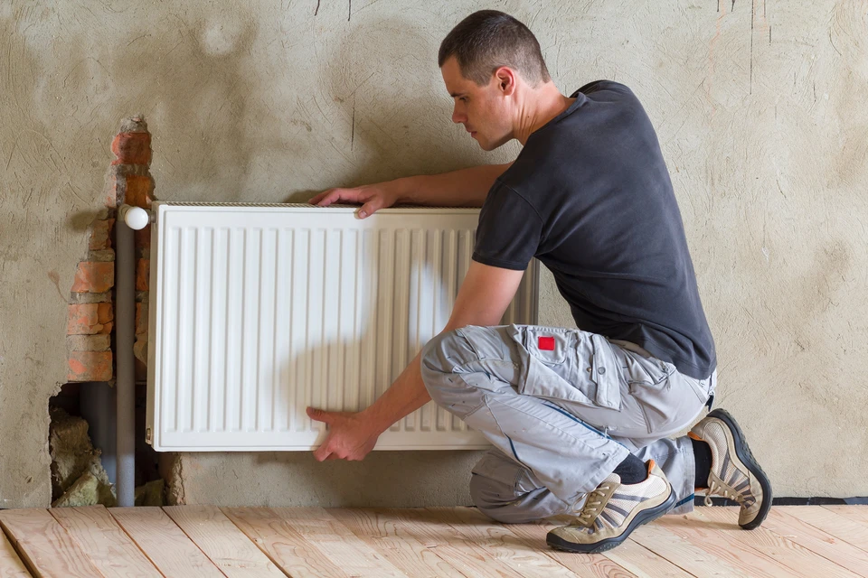 Un homme agenouillé installe un radiateur à panneau blanc contre un mur intérieur