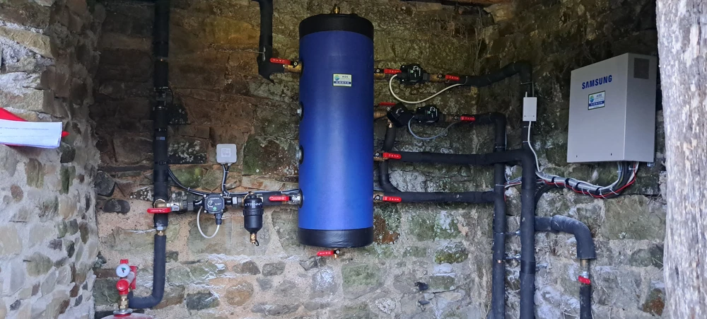 Unité extérieure de pompe à chaleur air/eau installée contre un mur en crépi beige, posée sur socle béton, entourée de végétation