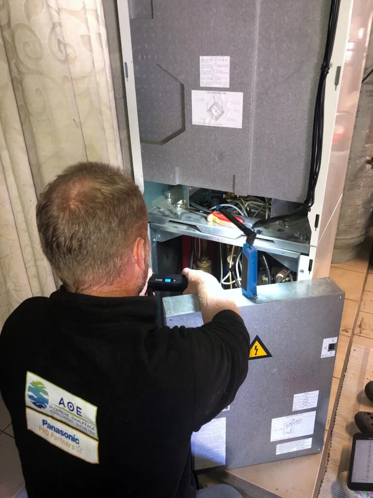 Un plombier en combinaison bleue allongé sur carrelage blanc sous un lavabo de salle de bain, tenant un tournevis, avec une caisse à outils contenant clés à molette, pinces et outillage spécialisé